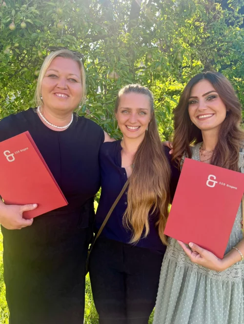 Gruppenfoto von drei Absolventinnen vor einem Birnenbaum. Zwei halten rote Abschlussmappen mit dem Titel SOB Bregenz