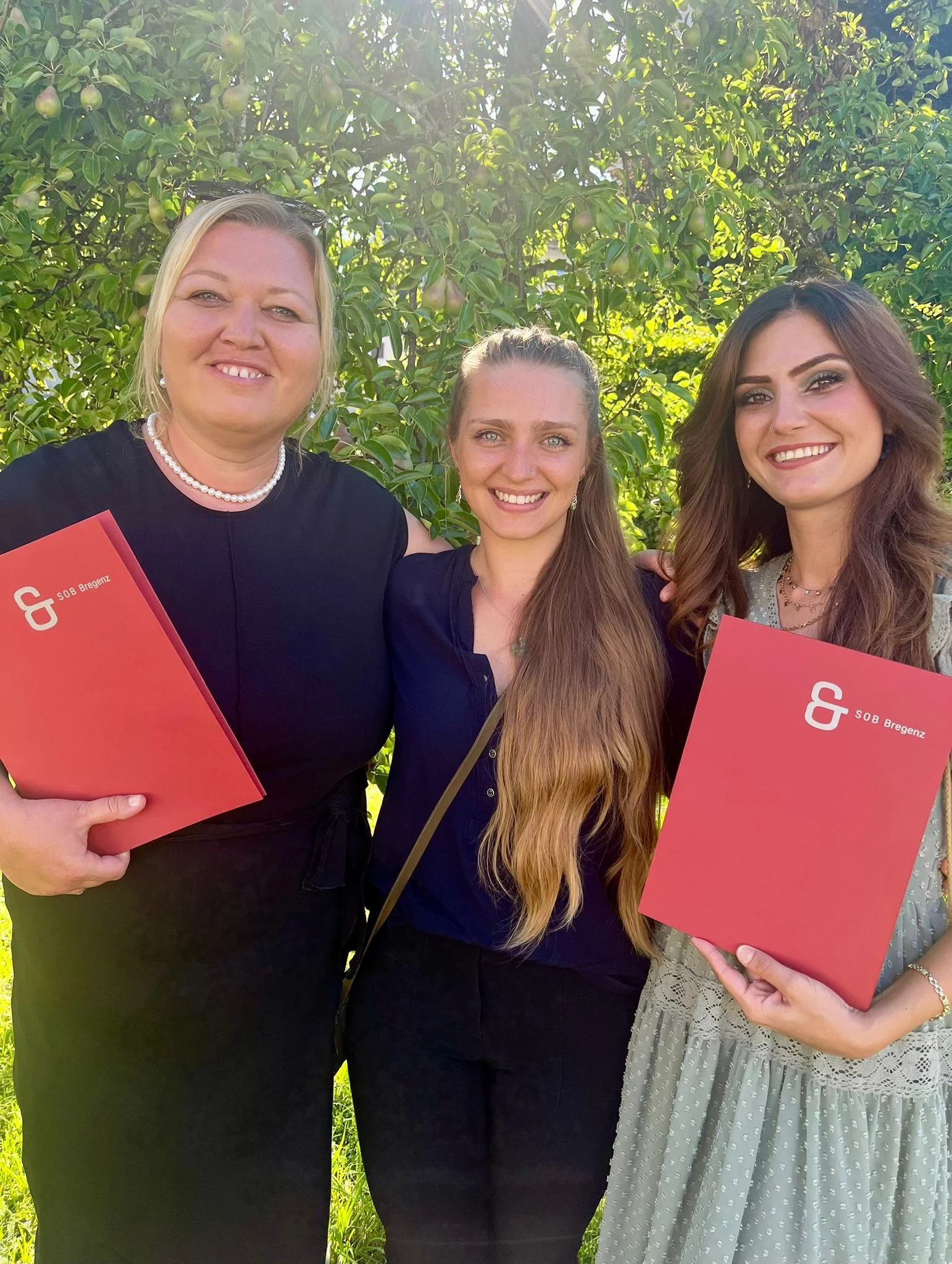 Gruppenfoto von drei Absolventinnen vor einem Birnenbaum. Zwei halten rote Abschlussmappen mit dem Titel SOB Bregenz